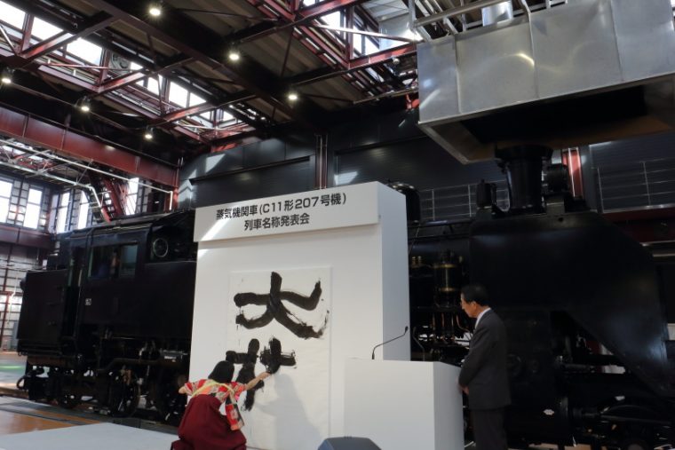 ↑南栗橋SL検修庫内で開かれた「列車名称発表会」。書道家・涼風花氏が「大樹」の字を描く