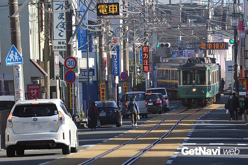 ↑通称「鎌倉市腰越電車通り」を走る20形電車。「電車接近」の表示に合わせ、車は道路脇に除けて電車の通過を待つ