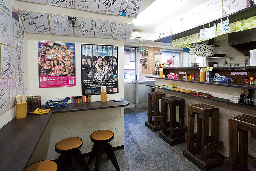 ↑店内 にはプロレス興行のポスターのほか、 サイン色紙が所狭しと貼られている。 有名レスラーのグッズを手掛ける黄 昏番長さんデザインの「宝そば」Tシ ャツは600枚売れた