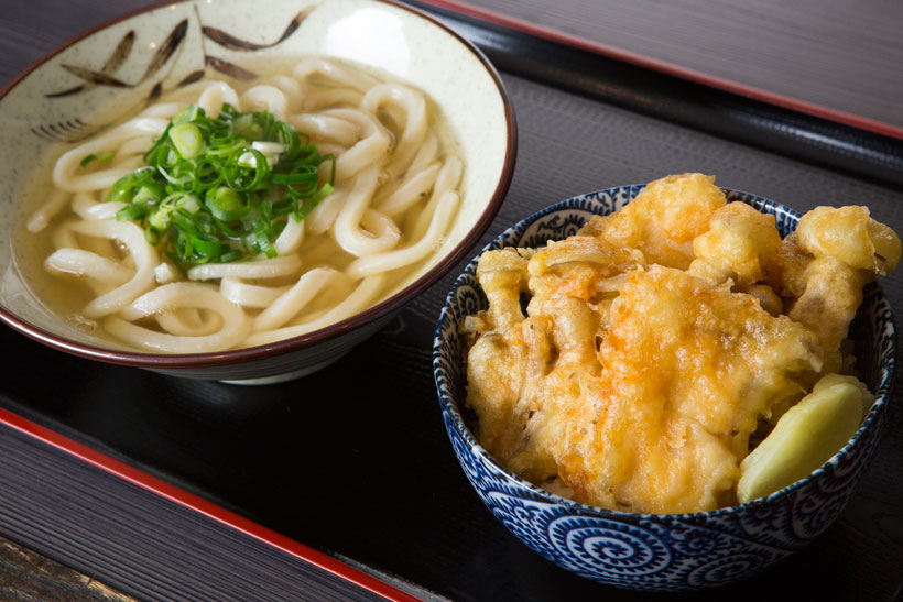 ↑きのこ天丼うどんセット（550 円）。．その日に仕入れた旬のきのこを2種類選んで天ぷらに。揚げたて天ぷらに染み込むタレのうまみが食欲を刺激する。かけうどんも付いて大満足