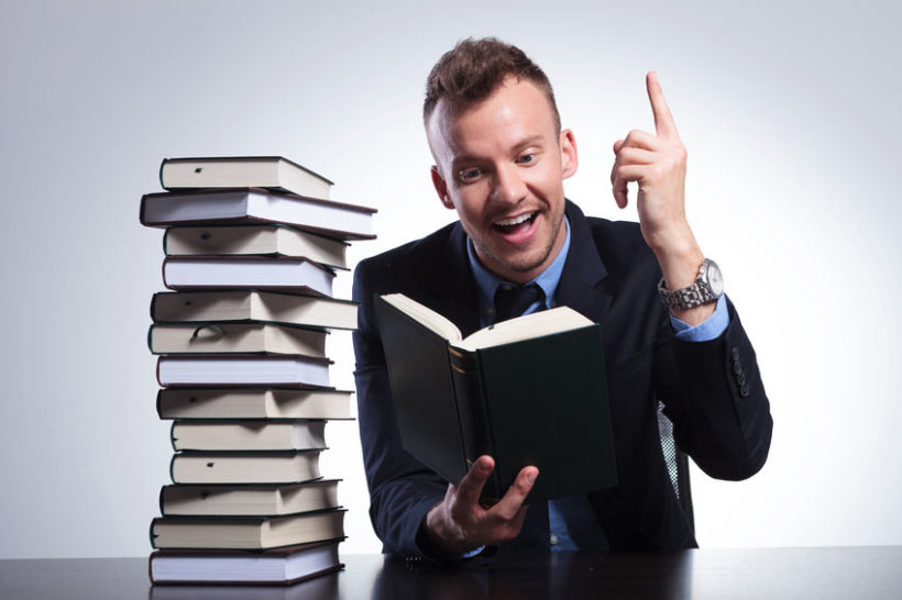 27838323 - young business man reading at an office with a stack of books and pointing up with a smile on his face, as he understands the idea. on a light studio background