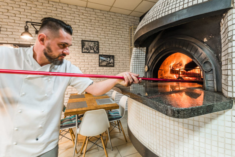 91210314 - handsome pizzaiolo man baking pizza in woodfired oven in local pizzeria